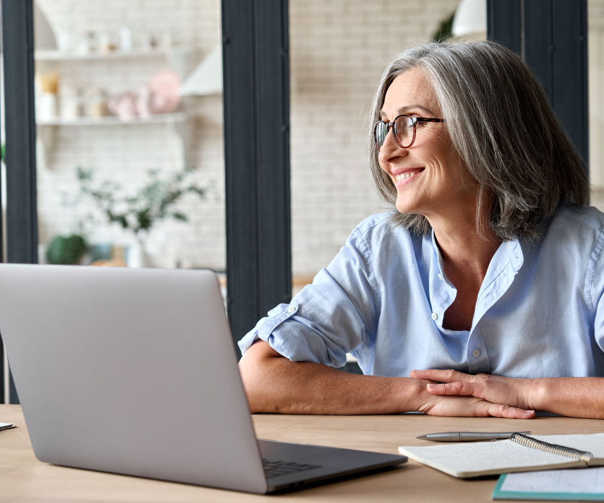 Joslin Rhodes - Middle-aged woman smiling in her kitchen with a laptop open in front of her, appearing relaxed and engaged.
