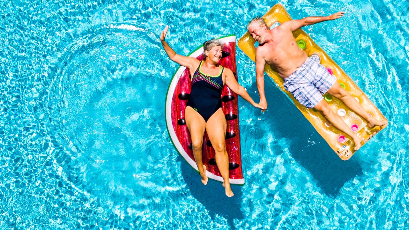 Retired couple relaxing on lilos in a large, sunny swimming pool – enjoying a peaceful holiday and the freedom of retirement.