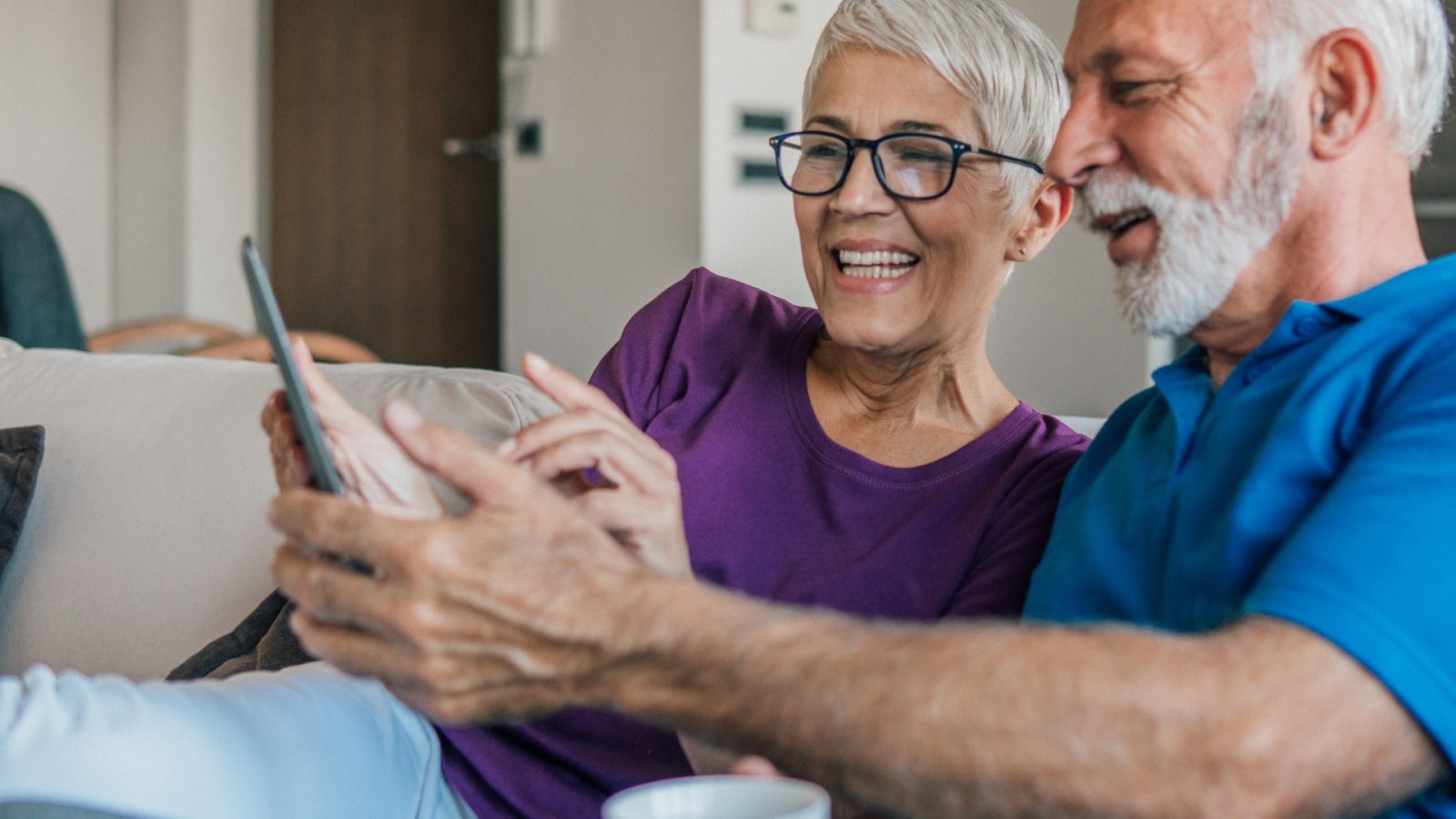 Joslin Rhodes - Image of a couple looking at an iPad smiling. Sat on the sofa in their