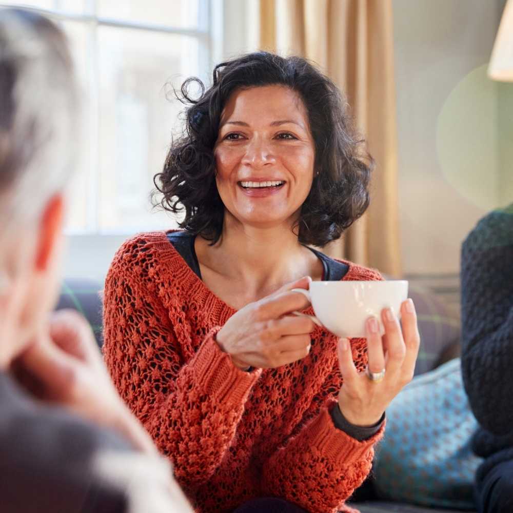 Joslin Rhodes – Woman wearing a red jumper in a living room, smiling and speaking to others while holding a coffee cup.