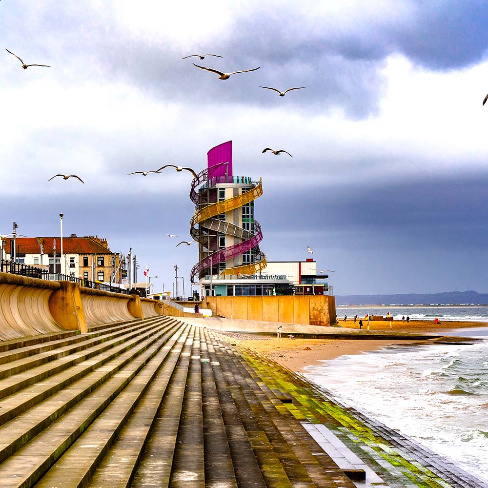 Joslin Rhodes – Redcar beach featuring the sea, a helter skelter amusement ride, and birds flying in the sky.