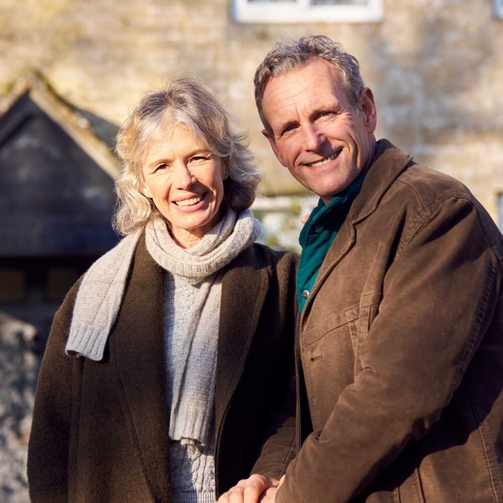 Joslin Rhodes - Retired man and woman smiling outside their house, dressed warmly in jackets and scarves.
