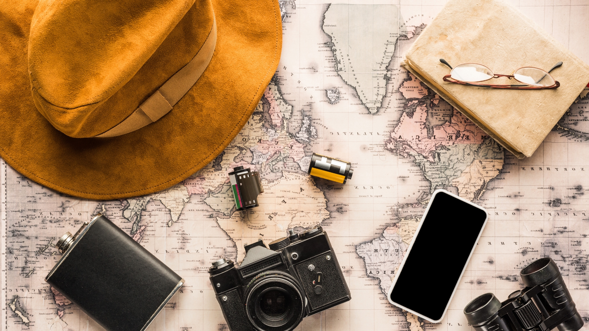 Flat lay image of travel items on a map background, including a hat, camera, phone, binoculars, glasses, and a notepad.