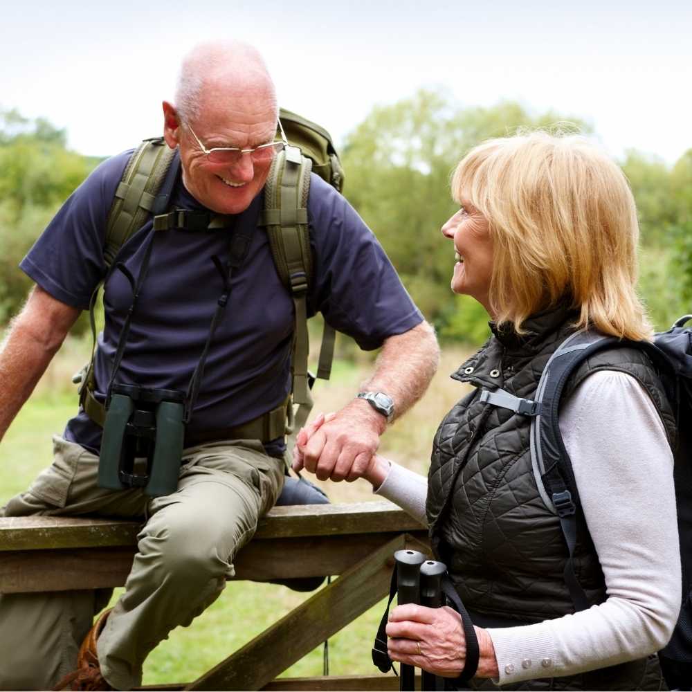 Joslin Rhodes – Two retired people smiling as they hike outdoors, climbing over a fence.