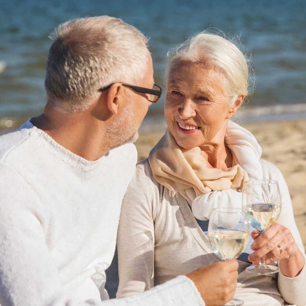 Joslin Rhodes - Middle-aged couple smiling at the beach, toasting with glasses of white wine.