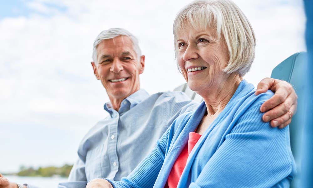 Joslin Rhodes - Happy retired couple outdoors. The man has his arm around the woman as they smile together. She is wearing a blue cardigan, and he is in a pale blue shirt.