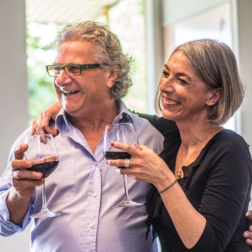 Joslin Rhodes - Joyful retired couple smiling and laughing as they celebrate with glasses of red wine, enjoying a relaxed and happy moment together.