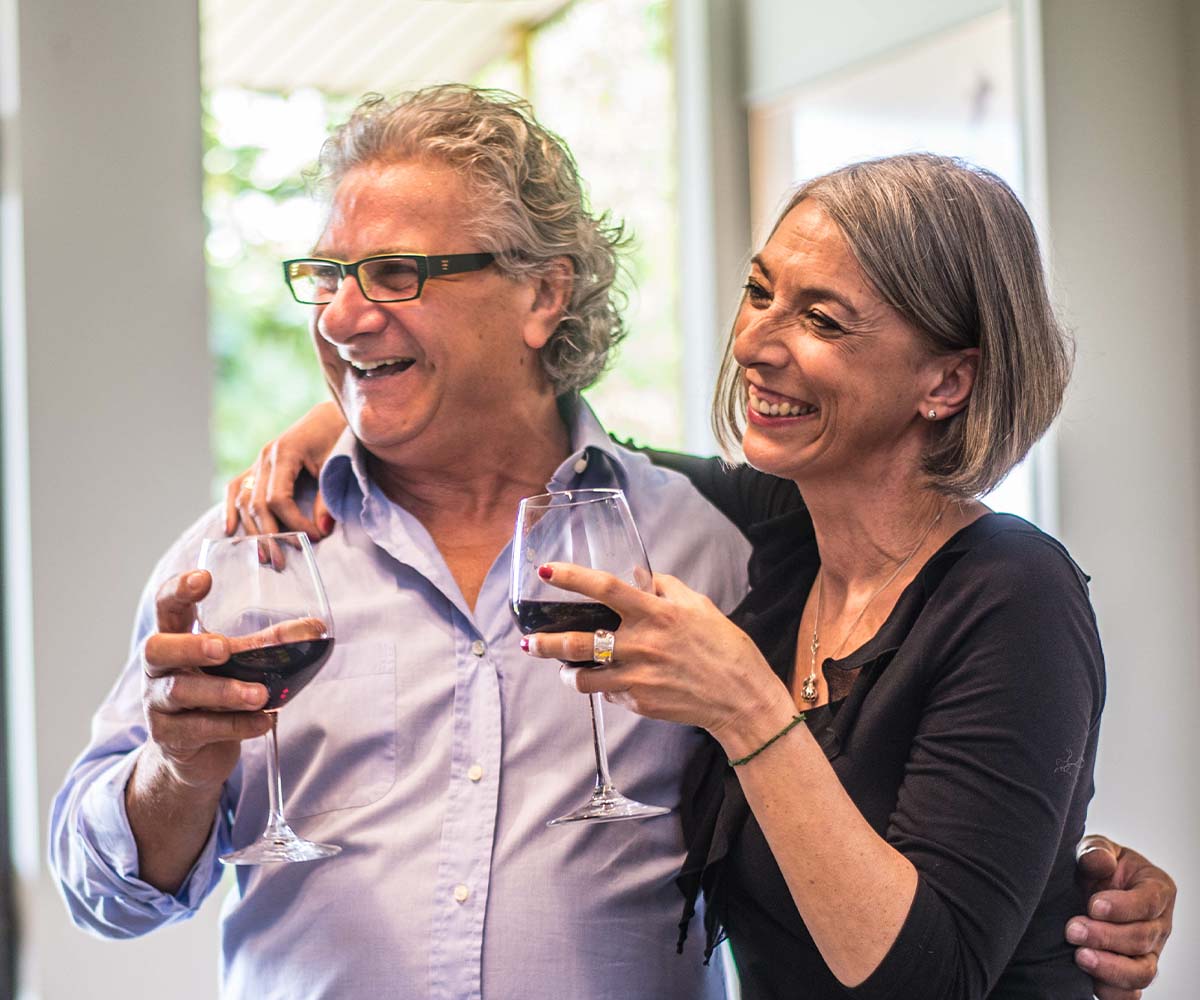 Joslin Rhodes - Joyful retired couple smiling and laughing as they celebrate with glasses of red wine, enjoying a relaxed and happy moment together.