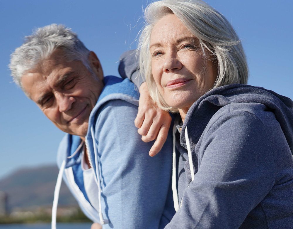 Joslin Rhodes - A middle-aged retired couple sitting closely together, smiling warmly as they enjoy a scenic view after a walk, embracing the joy of their retirement.