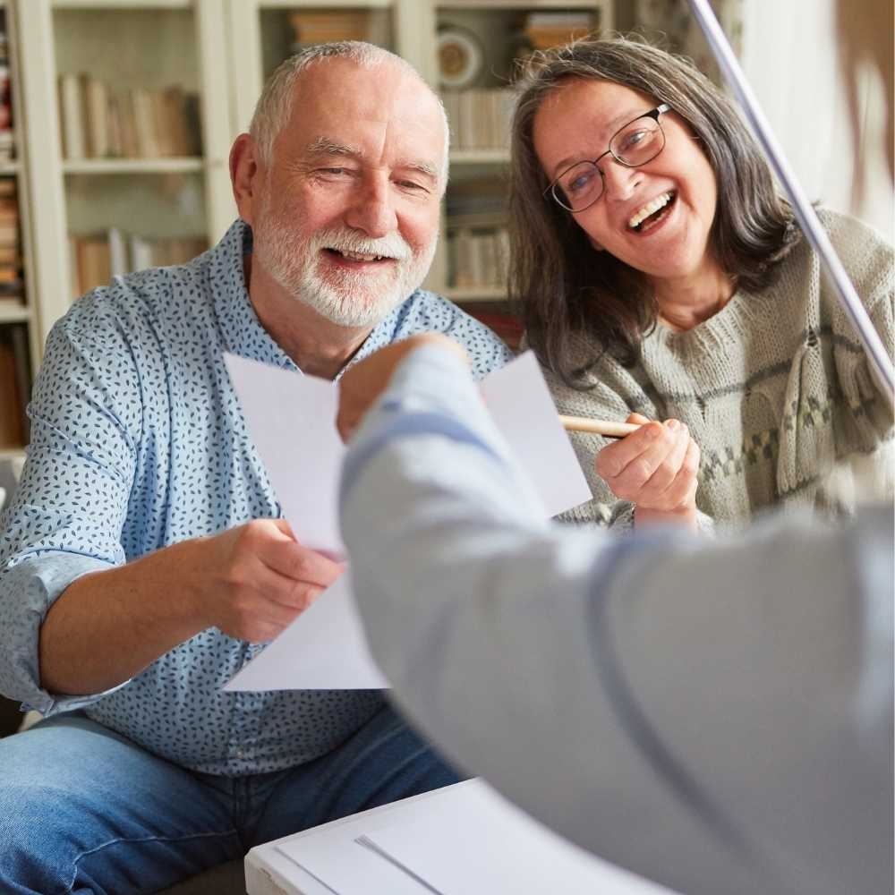 Joslin Rhodes - Middle-aged couple smiling in their living room as someone passes them a piece of paper.