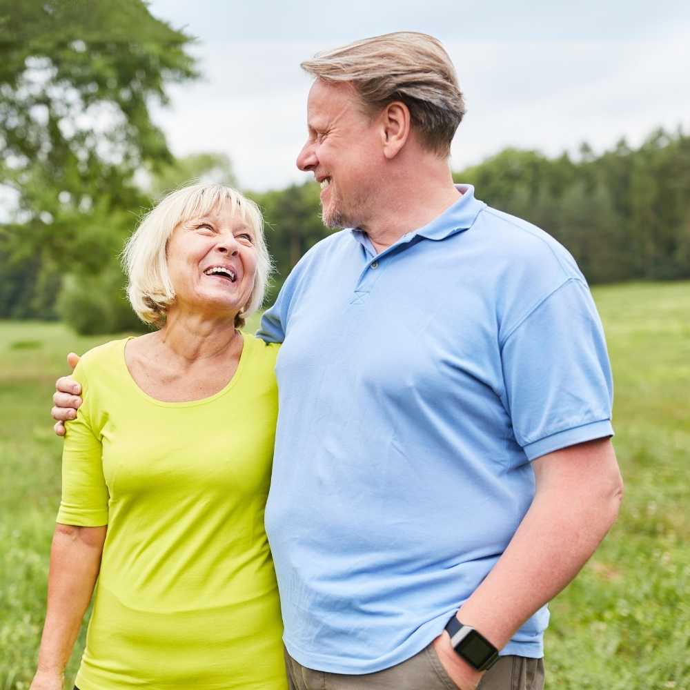 Joslin Rhodes - A retired couple standing outside, the woman wearing a lime green T-shirt and the man in a light blue shirt. They are looking at each other and laughing, with grass, a hedge, and a cloudy sky in the background.