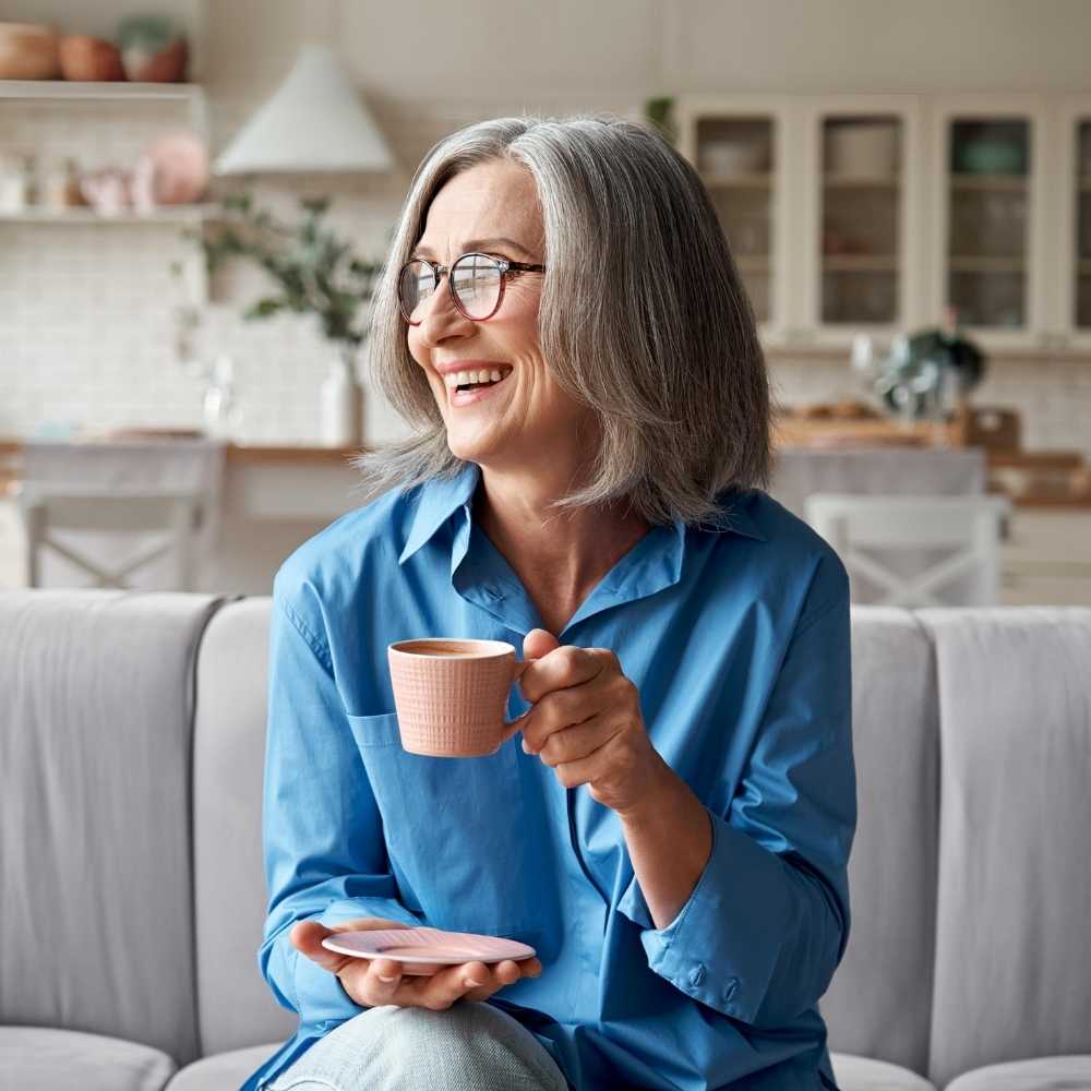 Joslin Rhodes - Smiling retired woman in a blue shirt, holding a cup of coffee and looking to the left while relaxing at home – capturing comfort and contentment.