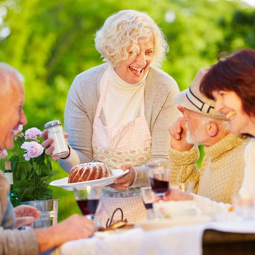 Joslin Rhodes - Group of retired people laughing and enjoying a sunny outdoor gathering, with a woman holding a cake who appears to be the host, surrounded by greenery.