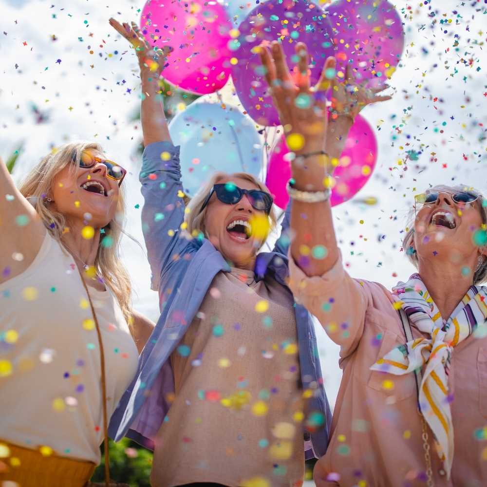 Joslin Rhodes - Retired people smiling and celebrating with arms raised, surrounded by purple, pink, and light blue balloons and confetti.