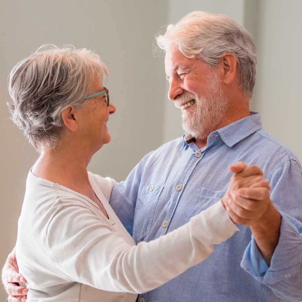 Joslin Rhodes - Retired couple smiling and looking at each other while dancing in their kitchen, enjoying a joyful moment together in retirement.